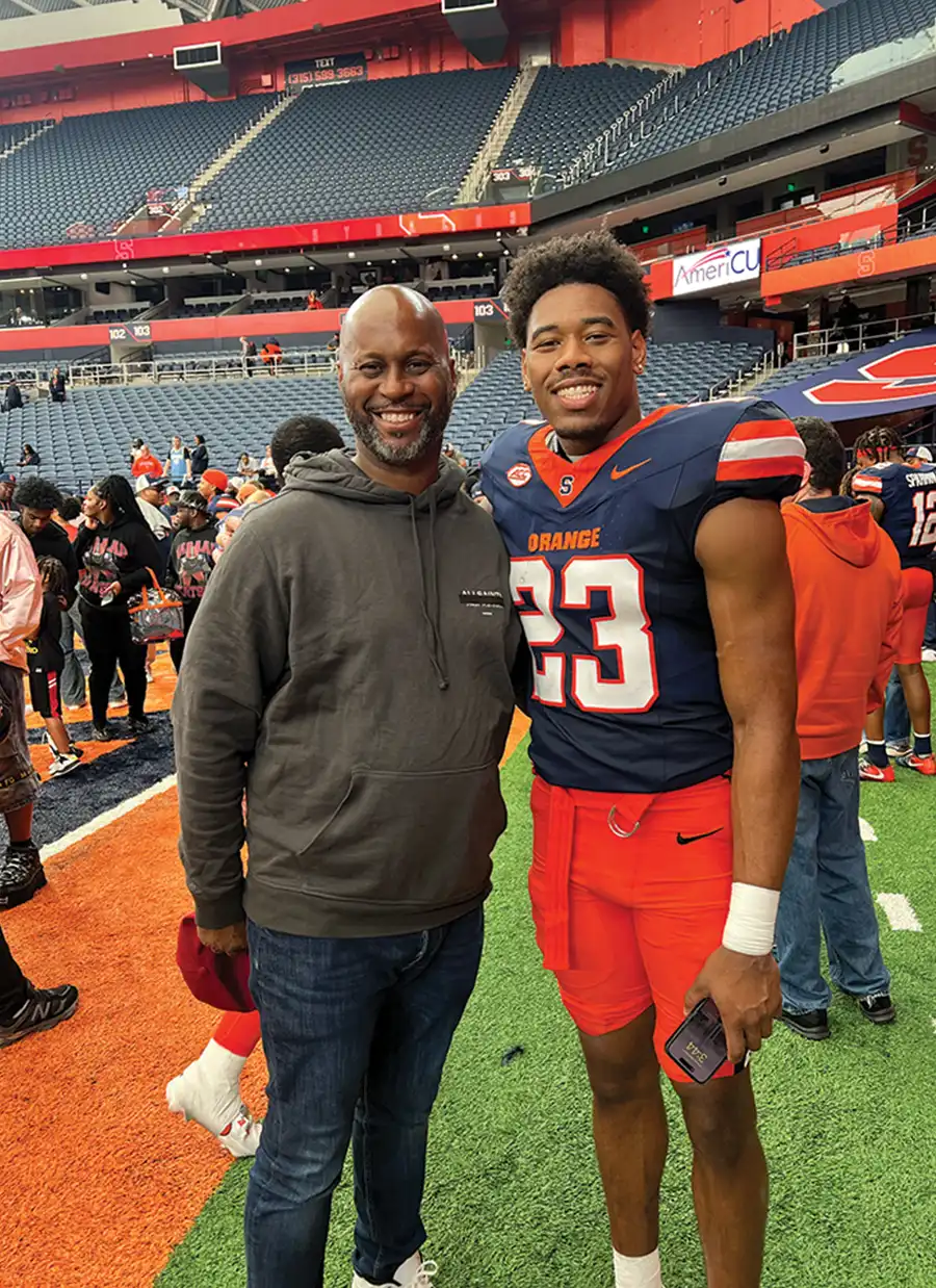 Ford stands on a football field next to Devin Grant. Grant wears a blue and orange Syracuse University jersey with the number 23 and orange shorts. Blue stadium seating is visible behind them.