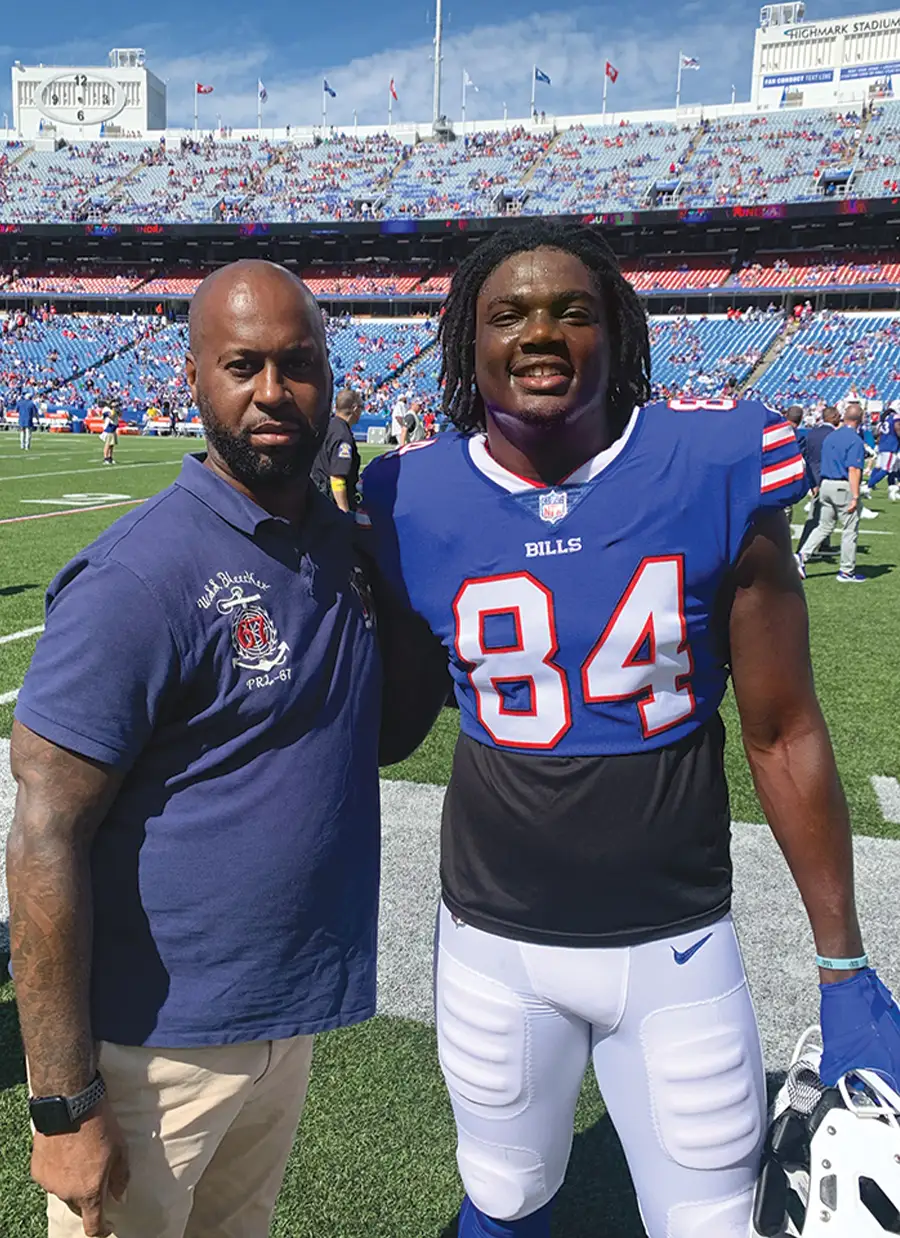 Ford stands on a sunny football field with Jalen Wydermyer. Wydermyer wears a blue Buffalo Bills jersey with the number 84 and white pants. A large crowd and stadium seating are in the background.
