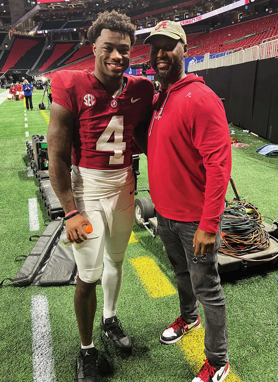 Ford stands with Jalen Milroe on a football field. Both wear red tops, with Milroe in a red University of Alabama jersey with the number 4. Coiled cables and stadium equipment are on the turf behind them.