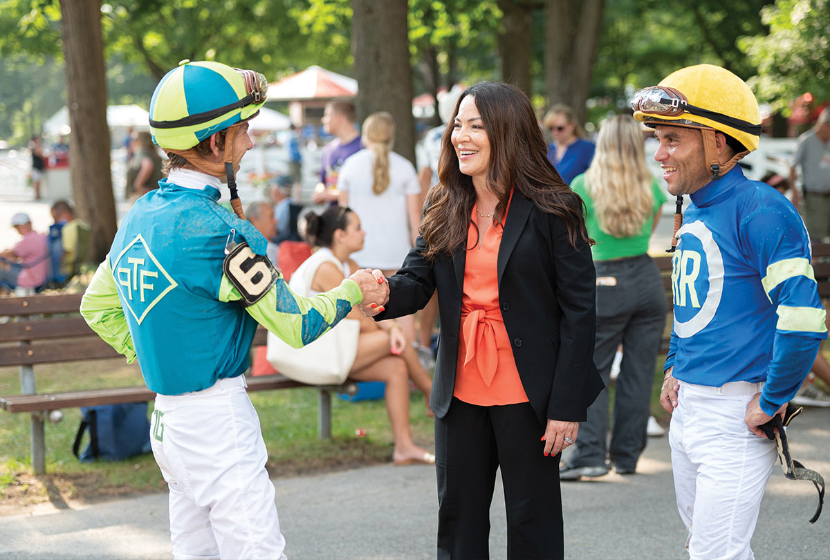 Lisa Lazarus, in a black blazer and orange top, shakes hands with jockey Tyler Gaffalione at the Saratoga Race Course. Jockey Joel Rosario stands to the right. Both jockeys wear colorful racing silks, helmets, and goggles.