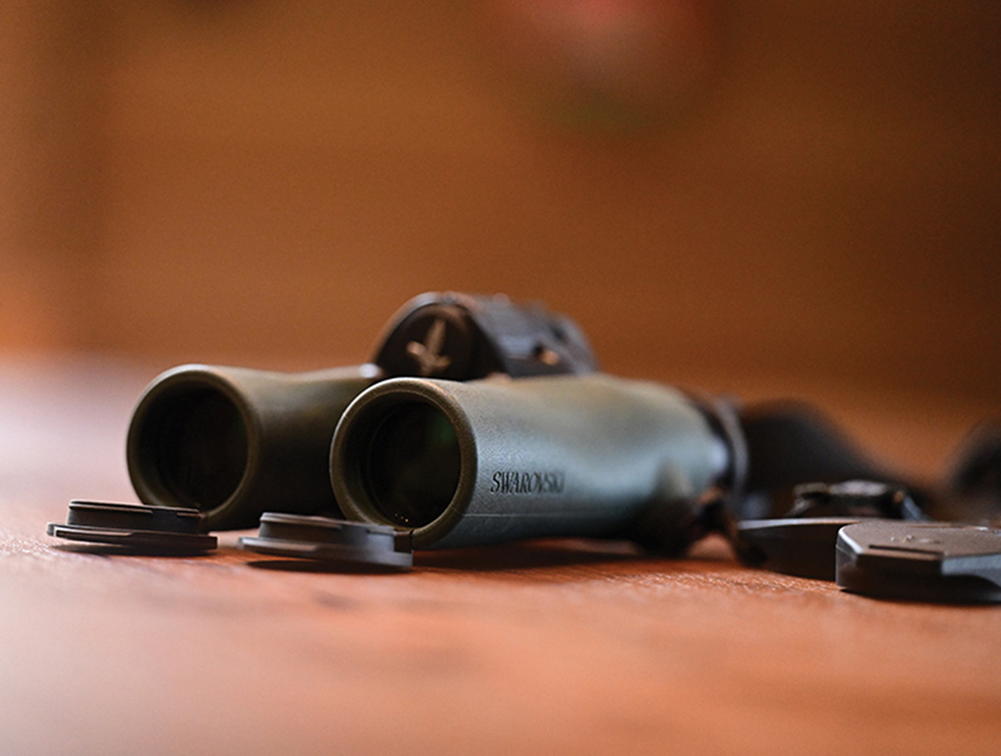 A close-up of green Swarovski binoculars resting on a wooden surface with the lens caps lying nearby.