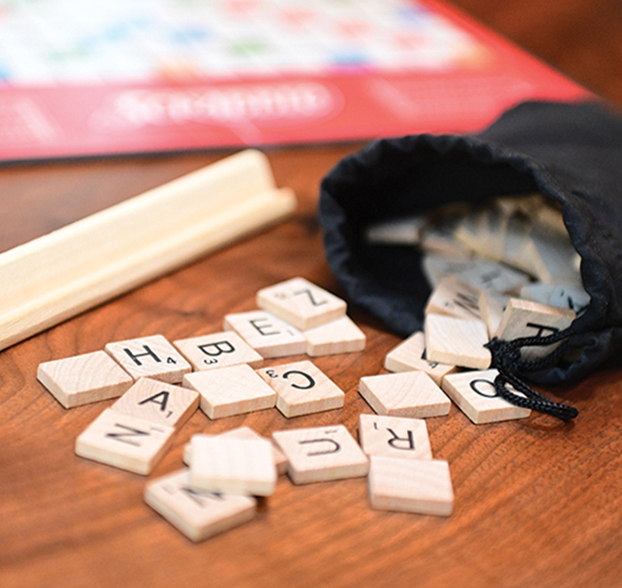Scrabble letter tiles spill out of a black fabric bag onto a wooden surface next to a wooden tile holder and a game board.