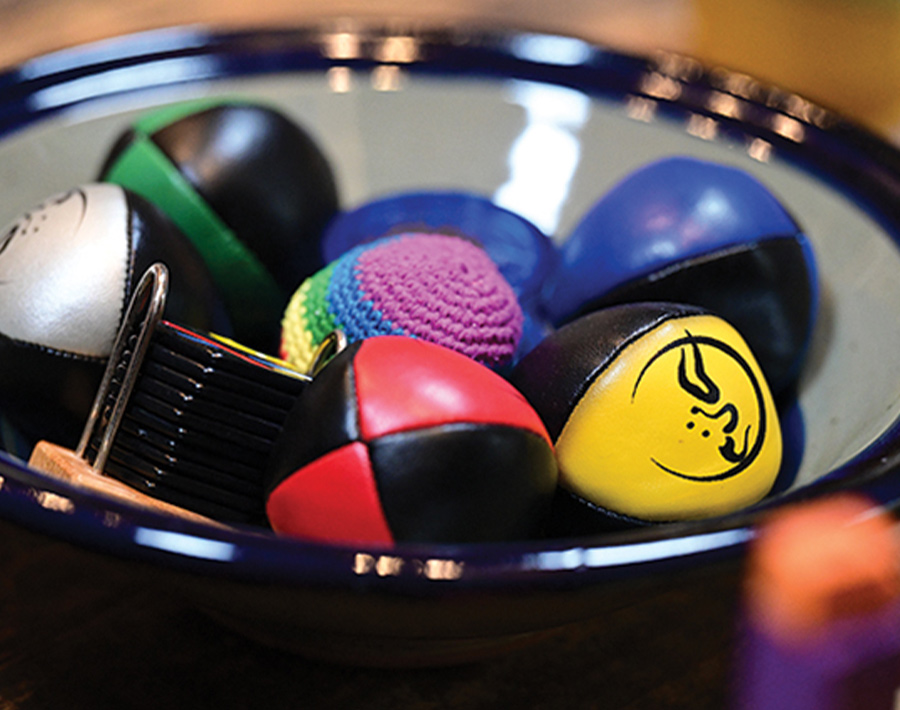 A close-up of a glass bowl containing several multi-colored juggling balls made of various leather and crochet materials.