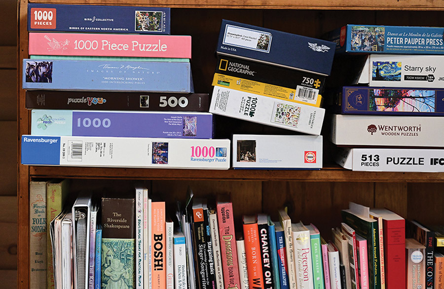 A wooden bookshelf stacked with numerous jigsaw puzzle boxes on the upper levels and a variety of books on the lower shelf.