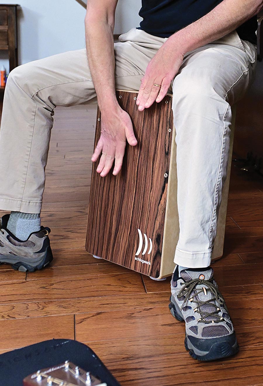 A close-up of Professor Howard Erichson sitting on and playing a wooden cajón drum with his hands. His legs frame the instrument on a hardwood floor.