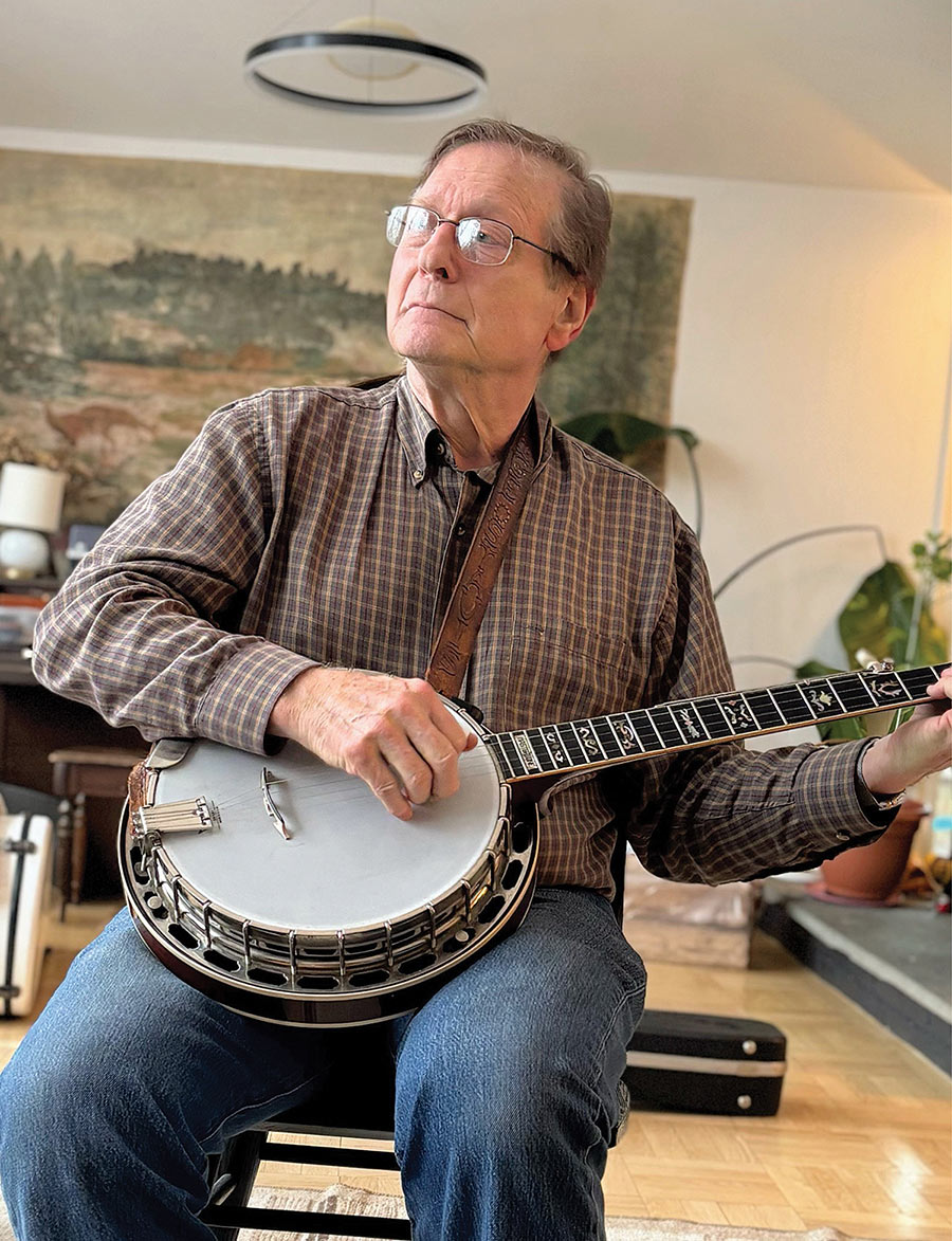 George Stavis sits and looks upward while playing a banjo with a leather strap.