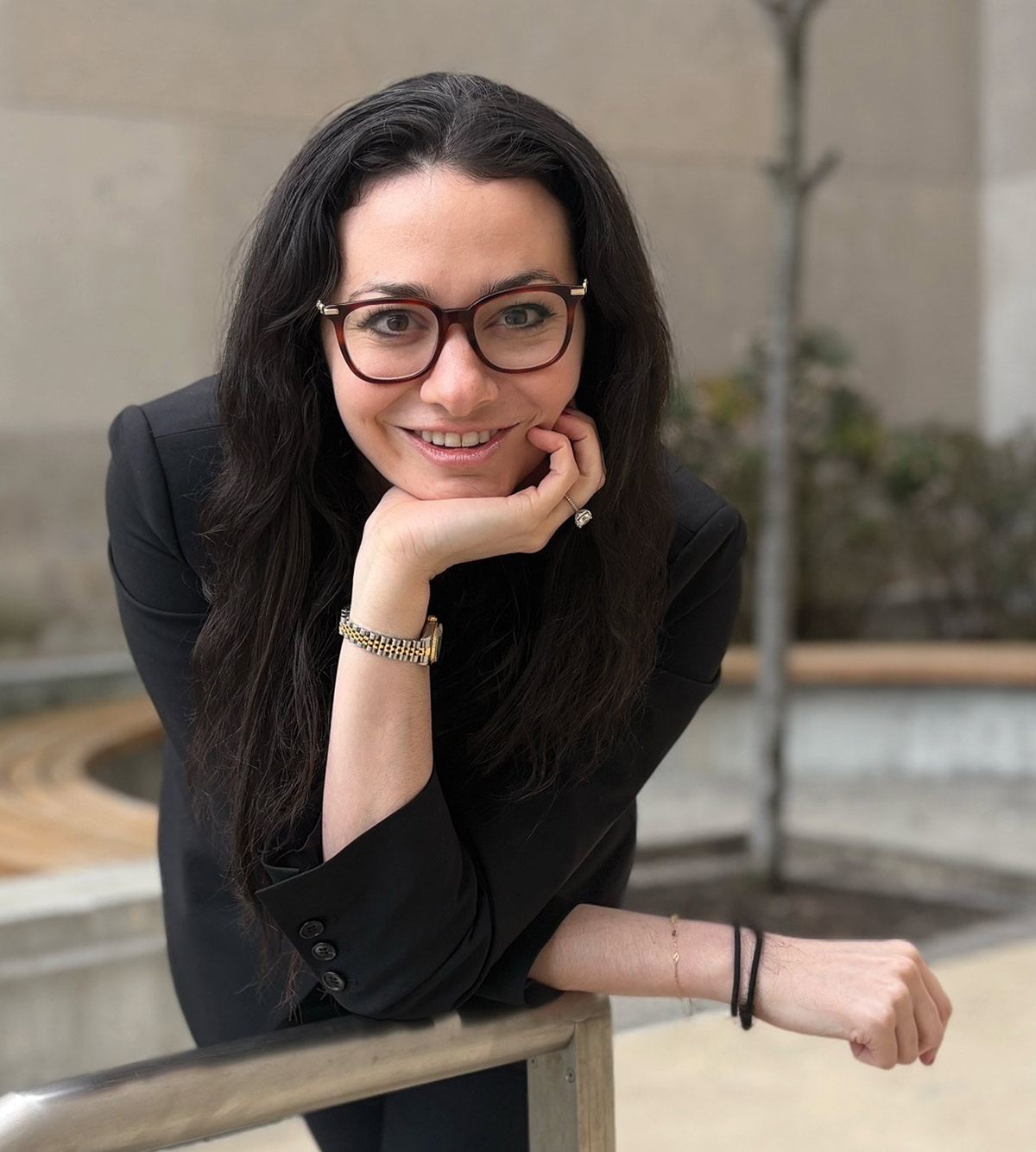 A smiling woman with long dark hair and glasses leaning on a metal railing, resting her chin on her hand while wearing a black suit jacket.